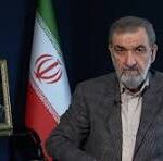 A man with a gray beard and blazer sits at a desk beside an Iranian flag and a framed portrait on a side table.