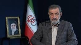 A man with a gray beard and blazer sits at a desk beside an Iranian flag and a framed portrait on a side table.
