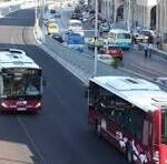 Two red city buses travel along a busy urban street with cars and pedestrians nearby.