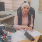 Woman wearing a pink hijab and vest sits at an office desk, reviewing papers with a tablet propped up in front of her.