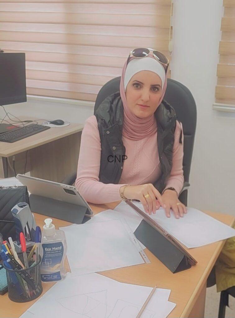 Woman in a pink hijab and vest sits at an office desk, reviewing papers with a tablet propped up beside her.