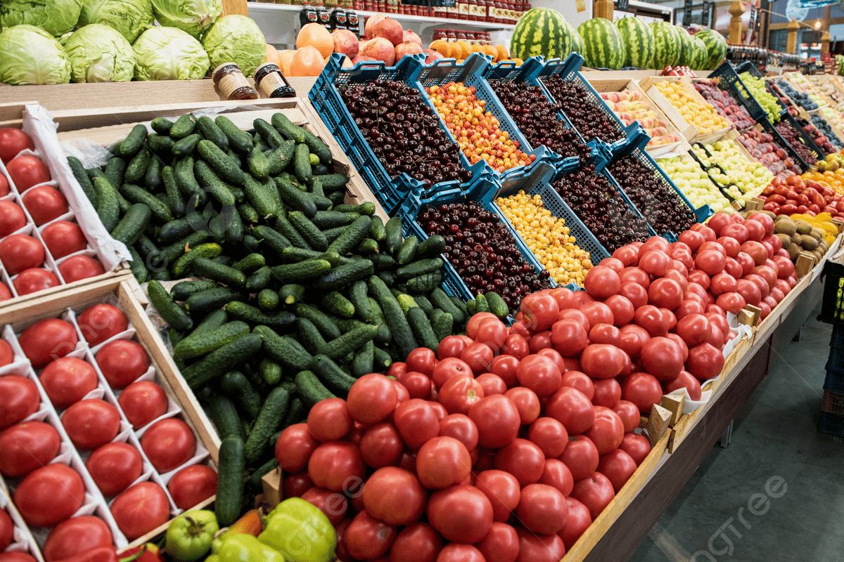 Fresh produce section with red tomatoes in the foreground, cucumbers piled beside them, and blue crates of small fruits in the middle at a supermarket.