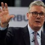 Man in a suit and tie with glasses raises his left hand while speaking at a public event or broadcast.