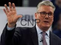 Man in a suit and tie with glasses raises his left hand while speaking at a public event or broadcast.