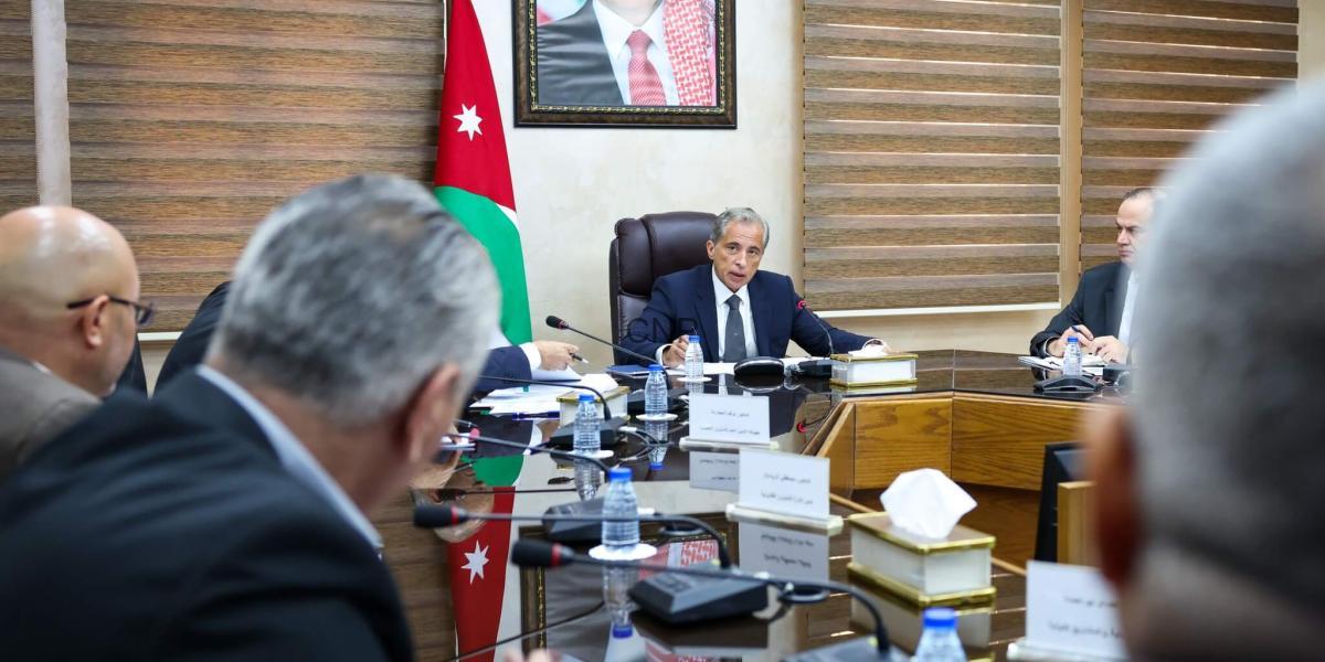 Man in a dark suit speaks at the head of a formal conference table as others listen, with a Jordan flag and portrait visible behind him.