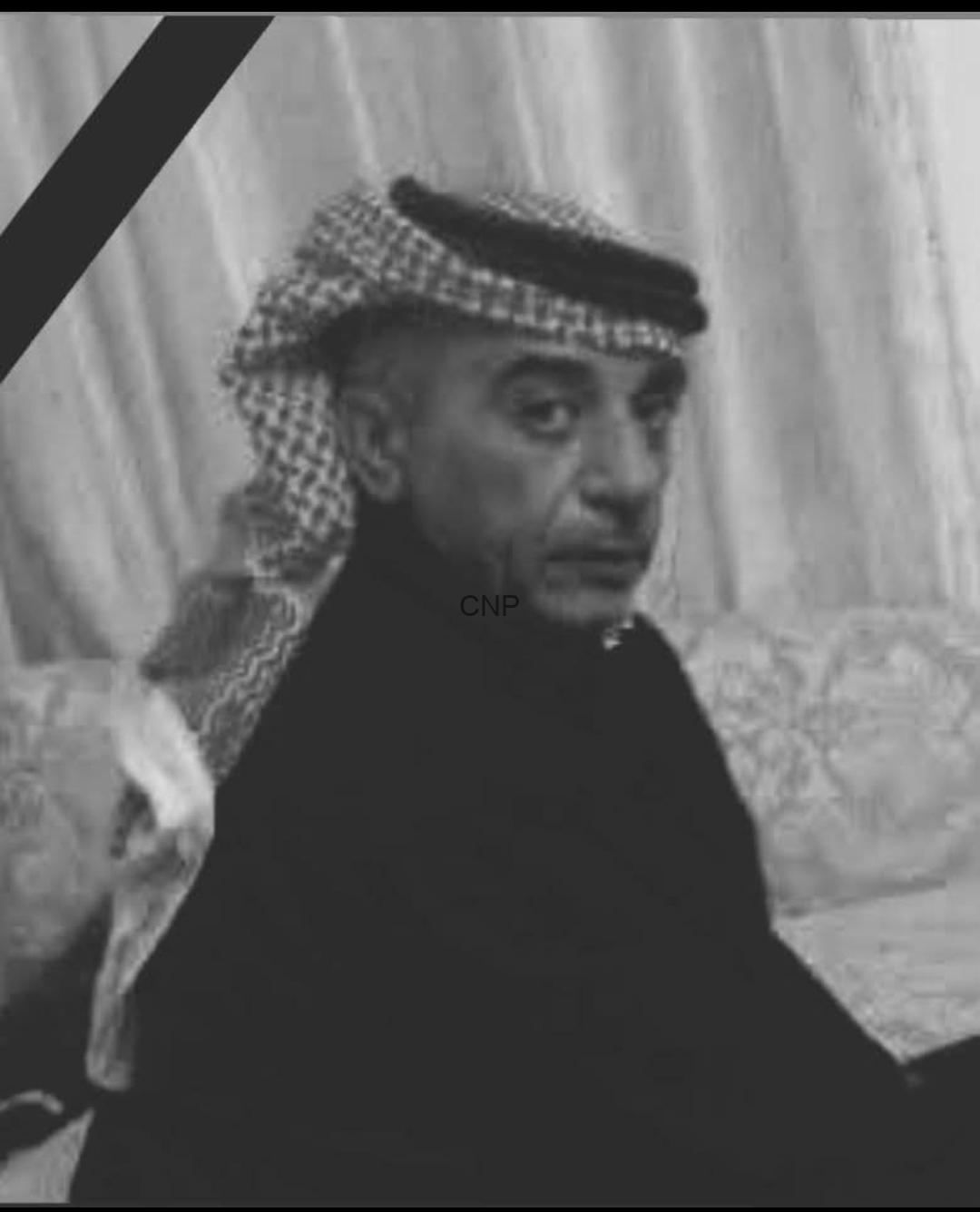 Black-and-white portrait of a man wearing a keffiyeh headscarf and dark clothing, looking toward the camera with a serious expression, mourning ribbon in the corner.