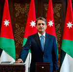 Man in a blue suit speaks at a podium with Jordan flags behind him in a formal government setting.【no punctuation】],