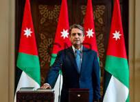 Man in a blue suit speaks at a podium with Jordan flags behind him in a formal government setting.【no punctuation】],