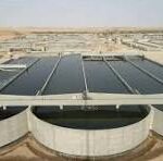 Rooftop solar panels on a low industrial building in a desert, with circular vent structures in the foreground and a blue sky above.