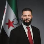 Portrait of a man with a beard in a dark suit and red tie, posed in front of the Algerian flag.