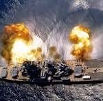 Warship on the water with two explosions and flames on its deck, producing smoke.