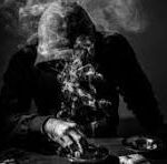 A person in a suit sits at a table in a dark, smoky scene with a skull in front of them.