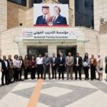 Group of about 25 people posing in front of a beige institutional building with an Arabic banner and a large poster of two men above the entrance, in a courtyard area with a patterned ground.