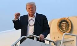 Man with white hair in a dark suit stands on airplane stairs, raising his right fist.