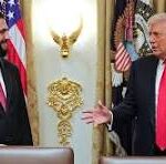 Two men in suits at a formal press event with American flags and a gold emblem in the background; one man gestures with open hands.