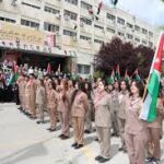Line of beige-clad students standing in formation, each holding a national flag during an outdoor ceremony outside a school building.