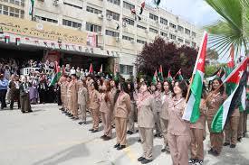 Line of beige-clad students standing in formation, each holding a national flag during an outdoor ceremony outside a school building.