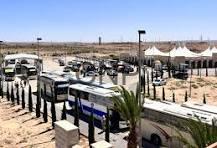 waha - Center News Network Line of tour buses and vans parked on a sandy desert lot, with a small palm plant in the foreground and tents in the background.