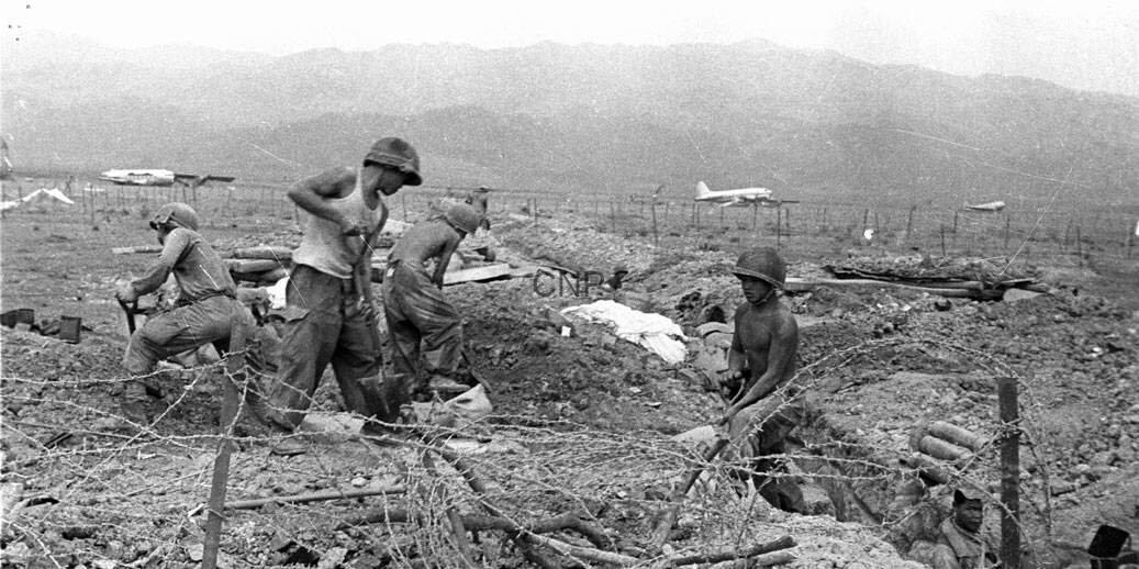 Soldiers digging in a trench with barbed wire in the foreground and a distant airplane in a barren landscape.