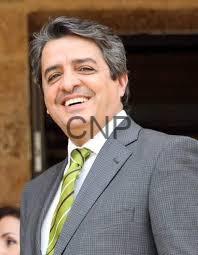 Smiling man in a gray suit with a green striped tie, standing outdoors near a brick structure.