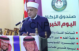 zzaa - Center News Network Man in a white traditional head covering speaks at a microphone from a podium at a charity event, with Arabic banners and flags in the background.