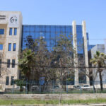 Modern office building with a beige stone left wing and blue glass center, fenced and lined with trees under a clear blue sky.