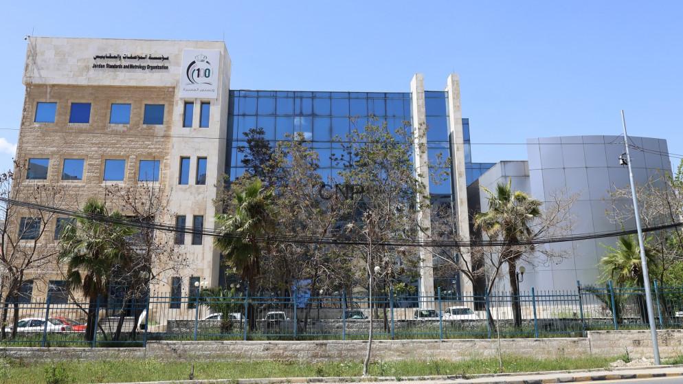 المواصفات والمقاييس - Center News Network Modern office building with a beige stone left wing and blue glass center, fenced and lined with trees under a clear blue sky.
