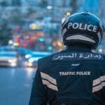 Traffic police officer seen from behind wearing a helmet and vest labeled 'Police' and 'Traffic Police' on a busy city street with flashing emergency lights in the distance