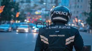 Traffic police officer seen from behind wearing a helmet and vest labeled 'Police' and 'Traffic Police' on a busy city street with flashing emergency lights in the distance