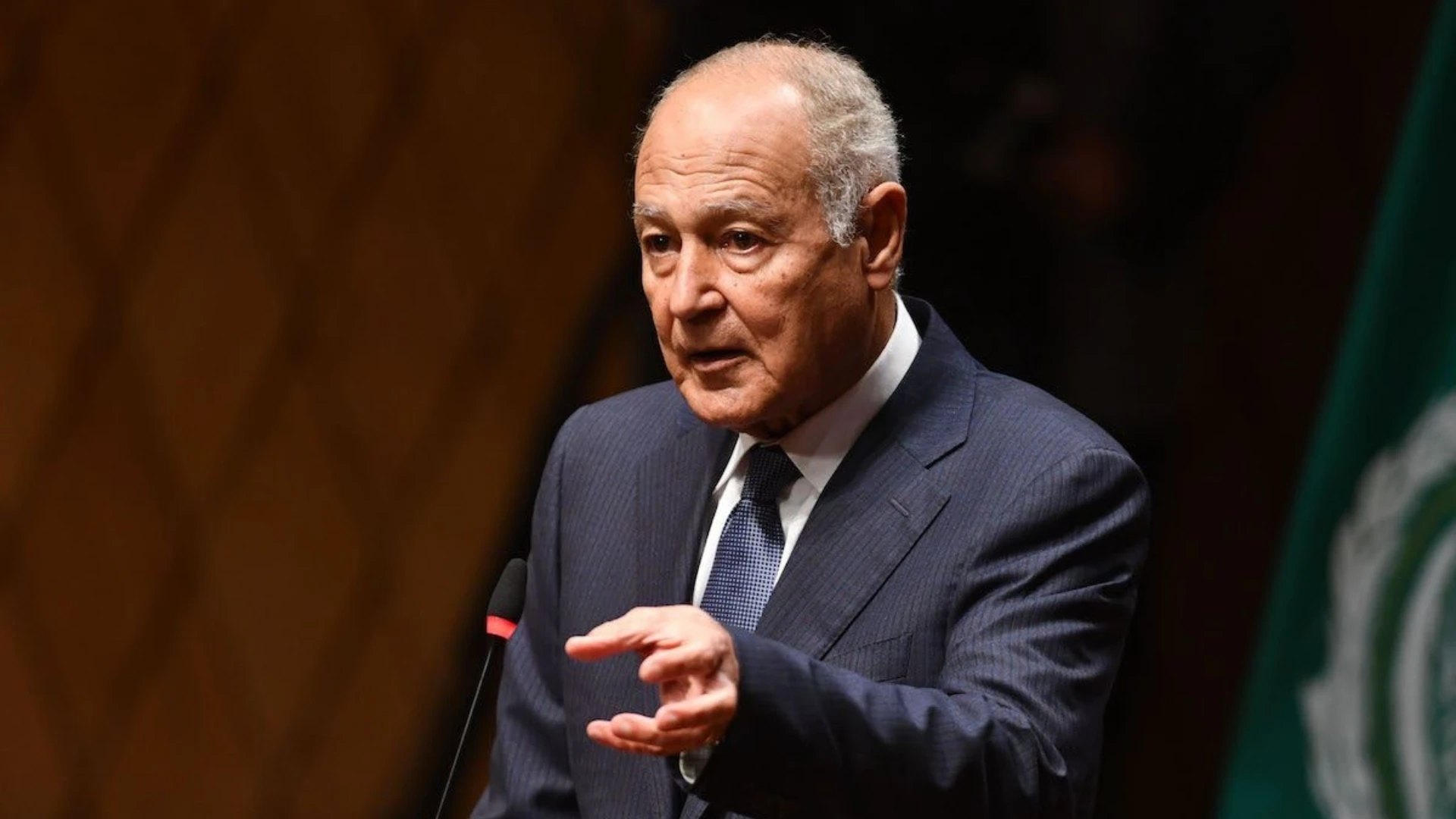 Older man in a dark pinstripe suit delivering a speech at a podium, gesturing with his hands.