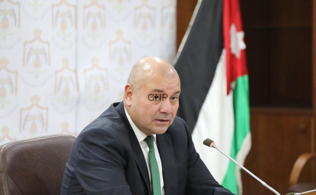 Bald man in a dark suit and green tie seated at a desk with a microphone, flag in the background.