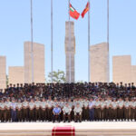Large group of uniformed military personnel posing for a formal group photo in front of a stone monument with flags flying overhead.