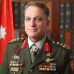 Portrait of a military officer in a green dress uniform with numerous medals and ribbons, standing in front of a bookshelf and a red flag.