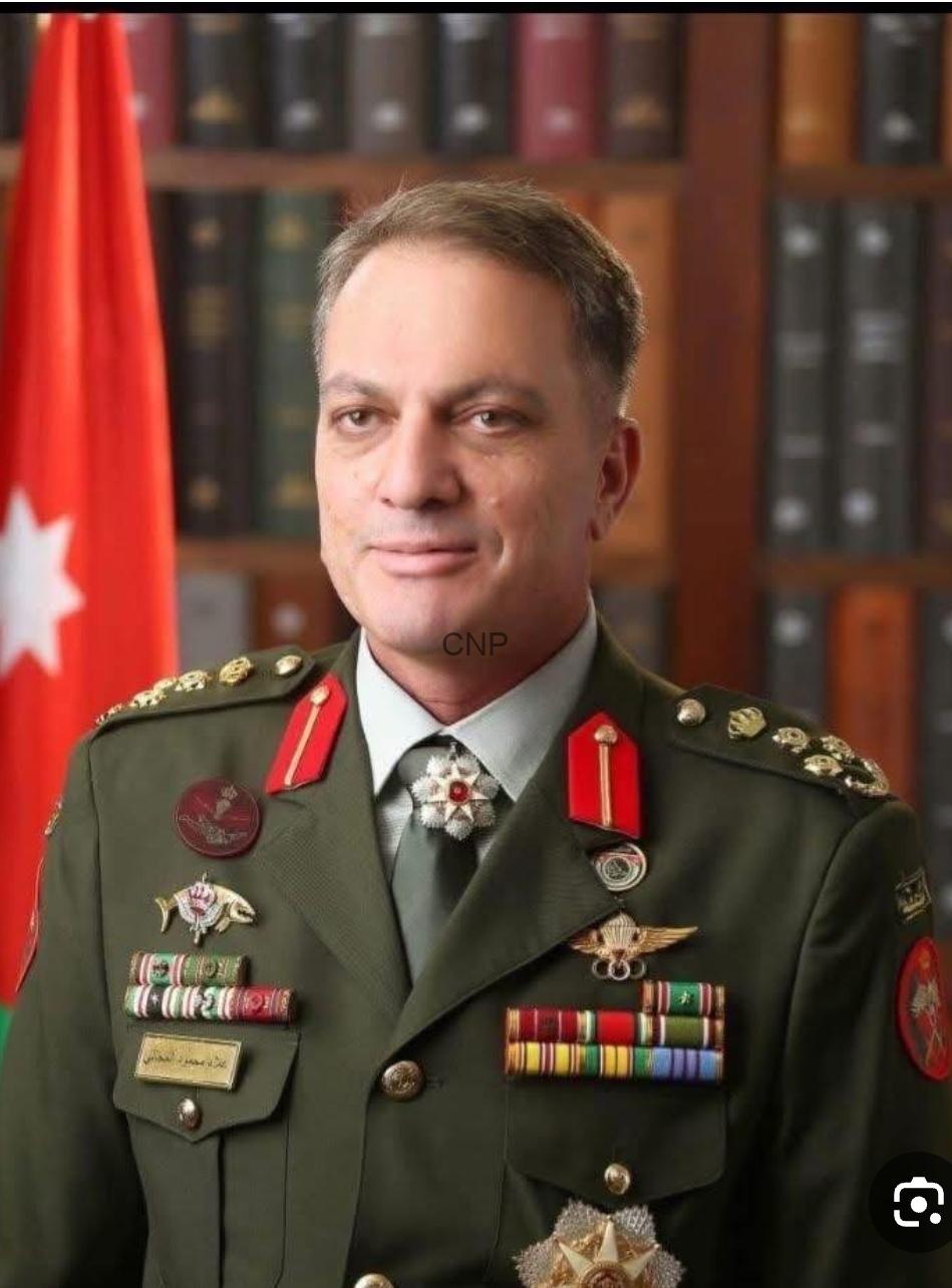 Portrait of a military officer in a green dress uniform with numerous medals and ribbons, standing in front of a bookshelf and a red flag.