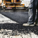 Road construction scene: worker in boots guiding asphalt being laid by a paving machine on a paved surface.