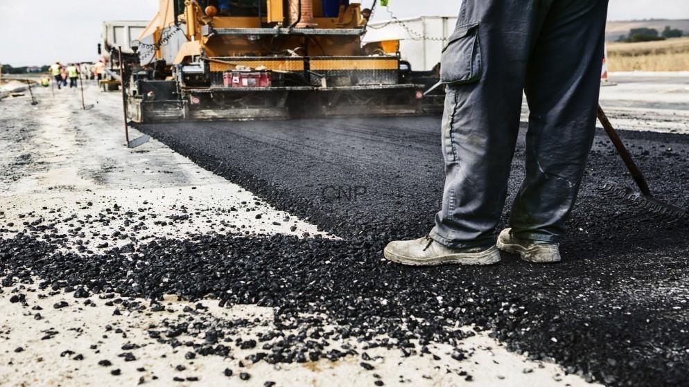 Road construction scene: worker in boots guiding asphalt being laid by a paving machine on a paved surface.