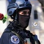 Tactical police officer in helmet and balaclava with a red rose near the shoulder, contrasting force and gentleness as a symbolic moment.