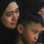 Mother in a black hijab consoling a crying young boy, with another woman in a hijab nearby.