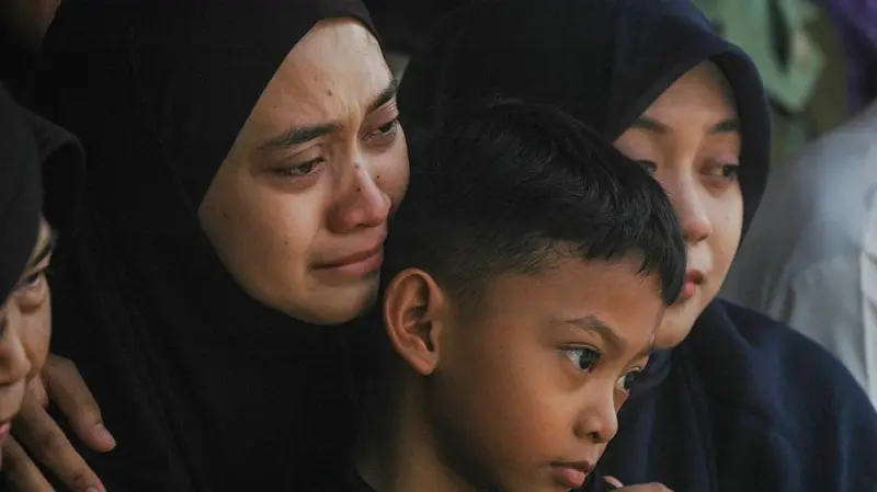 Mother in a black hijab consoling a crying young boy, with another woman in a hijab nearby.