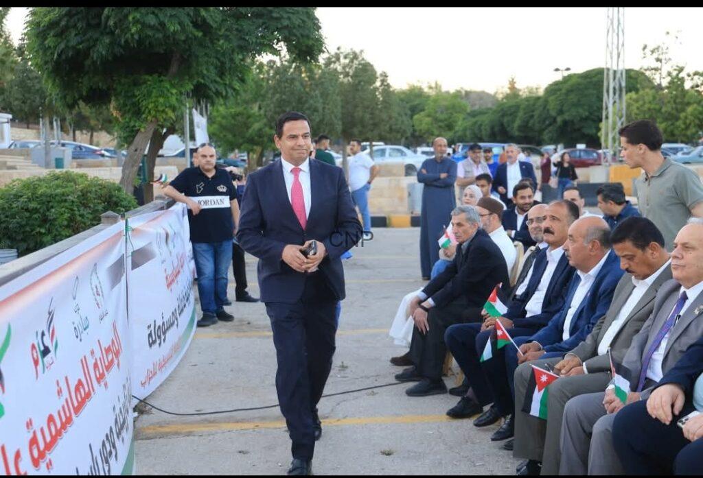 Man in a dark suit and red tie walks past a row of seated dignitaries holding Jordanian flags at an outdoor event, with banners along a railing.