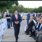 Man in a dark suit and red tie walks past a row of seated dignitaries holding Jordanian flags at an outdoor event, with banners along a railing.