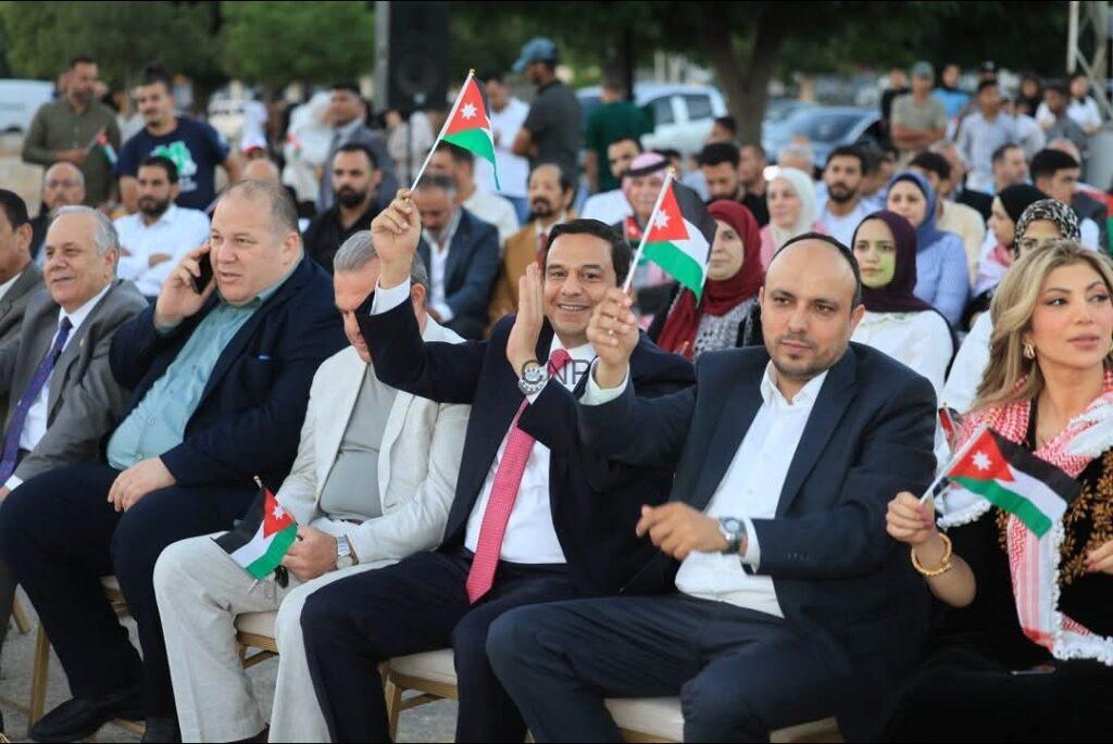 ayha2 - Center News Network Front row of an outdoor ceremony with men in suits waving Jordanian flags; crowd seated behind them looks on.