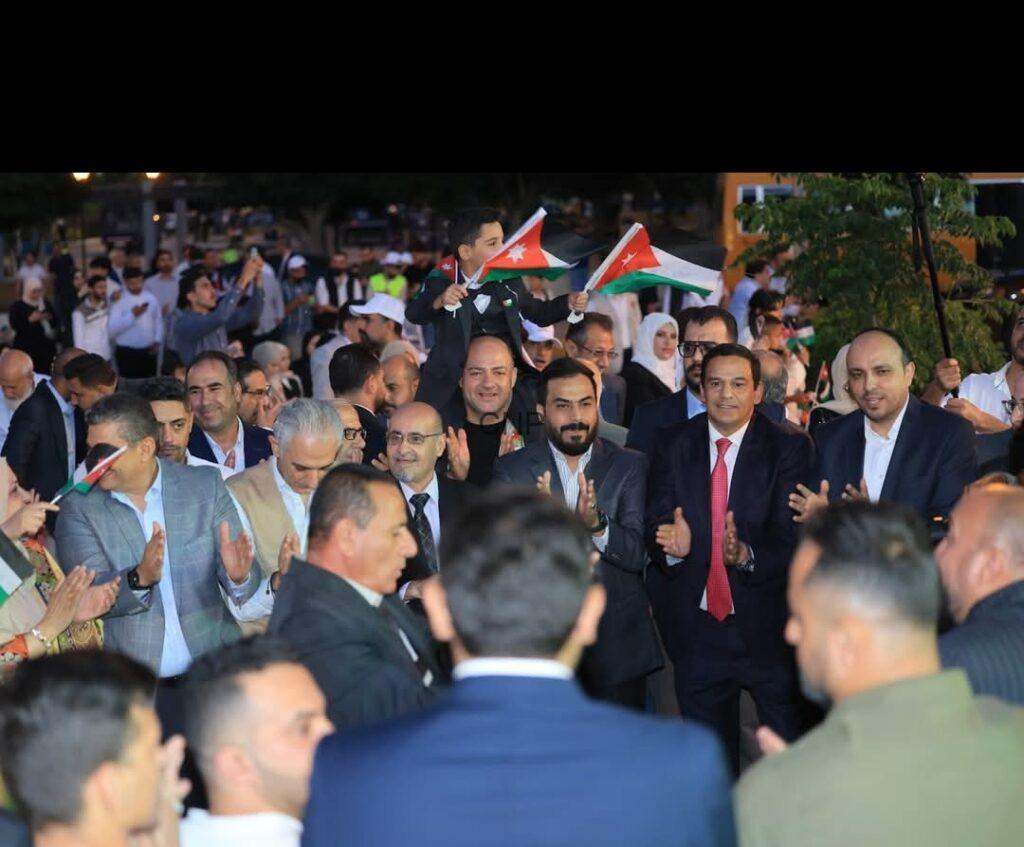 Crowd of well-dressed people at an outdoor rally, many clapping and waving Jordanian flags.