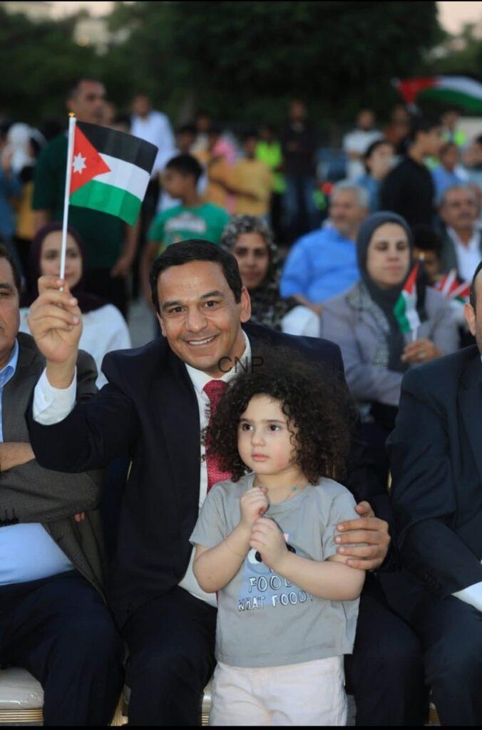 Smiling man in a suit waving a Jordan flag, seated with a curly-haired child at a crowded outdoor event.