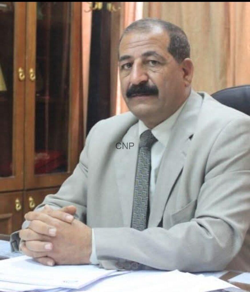 Professional man in a light gray suit and tie sits at a desk with folded hands in an office setting. Bookshelves are visible in the background.
