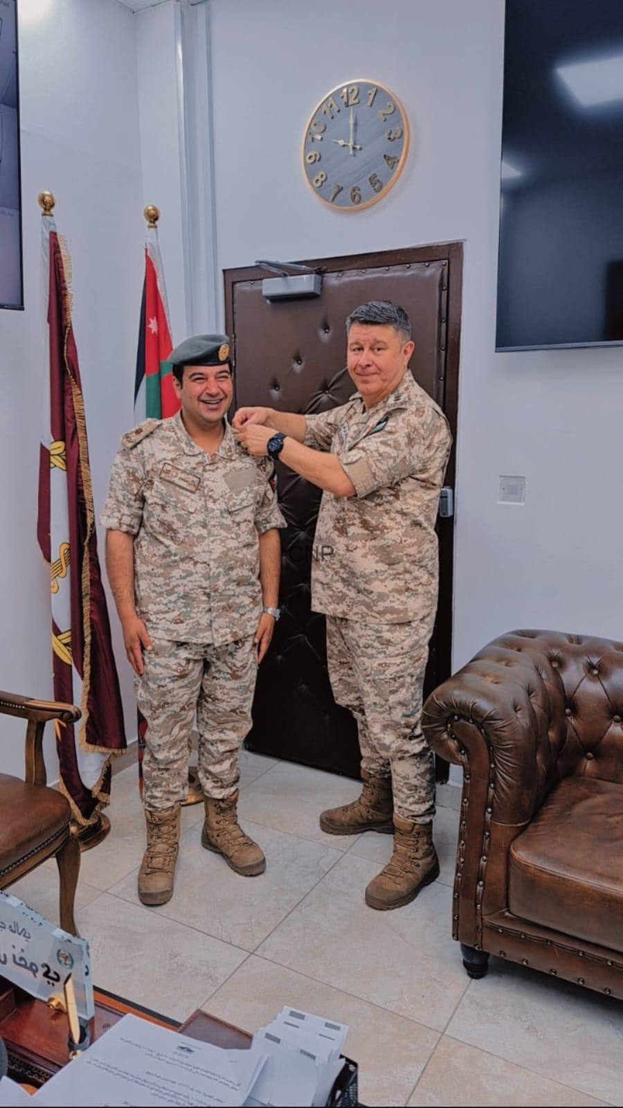 Two soldiers in desert camouflage stand in an office as one pins a medal on the other's chest, with Jordan’s flag and a second flag in the corner and a clock on the wall behind them.