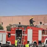Red fire truck with firefighters on the roof and a ladder extended toward a tan building under a clear blue sky, depicting an emergency response.