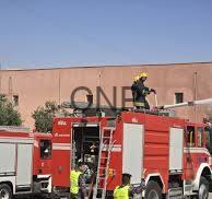 Red fire truck with firefighters on the roof and a ladder extended toward a tan building under a clear blue sky, depicting an emergency response.