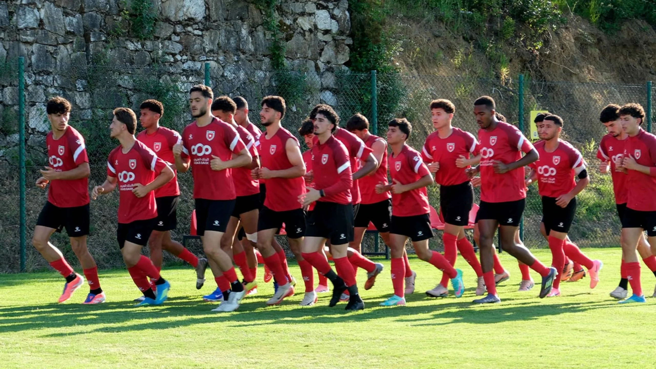 Group of young soccer players in red uniforms running on a sunny grass field during training.