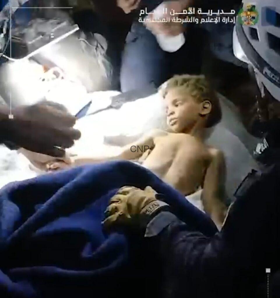 fan4 - Center News Network Boy lying shirtless on a hospital bed while medical staff attend to him in an emergency setting, bright light overhead.